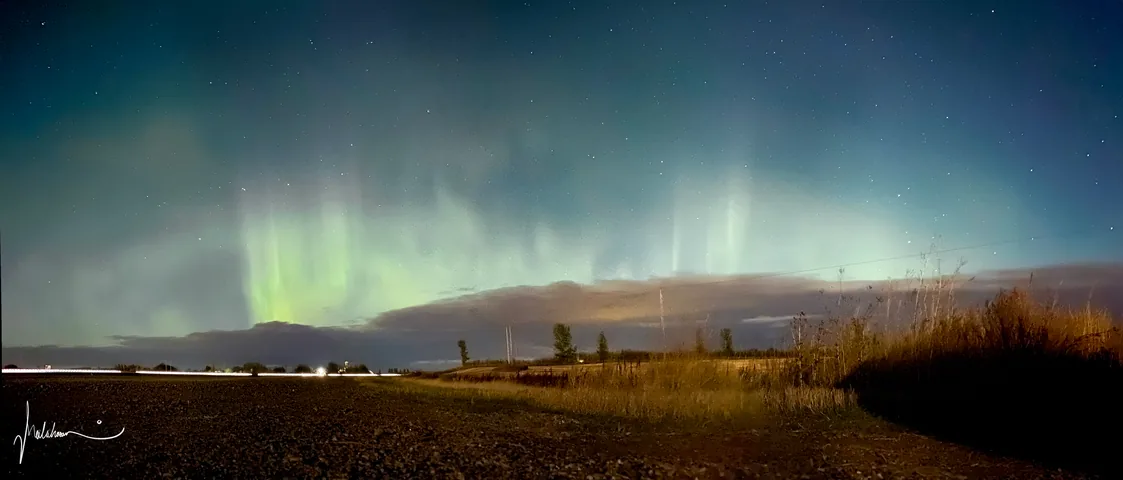 A panorama of green aurora pillars over a flat field with grass in the foreground.