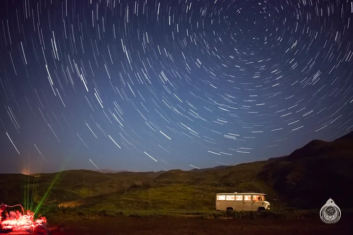 Star trails over hills, with a parked van and red-lit tents in the foreground.