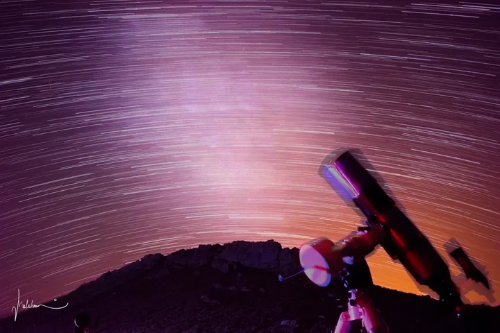 Long star trails streaking across a purple sky behind a telescope on its mount.